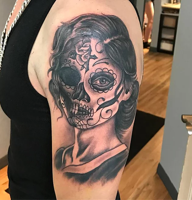 A tattoo of a sugar skull with half being a skull and half being a living face. 