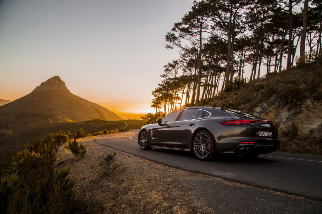 A luxury car drives into the sunset around the curve of a mountain. 