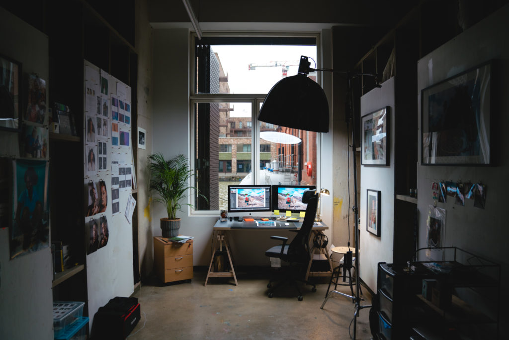 A dimly light office with  small desk and computer and many photos hanging on the walls. 