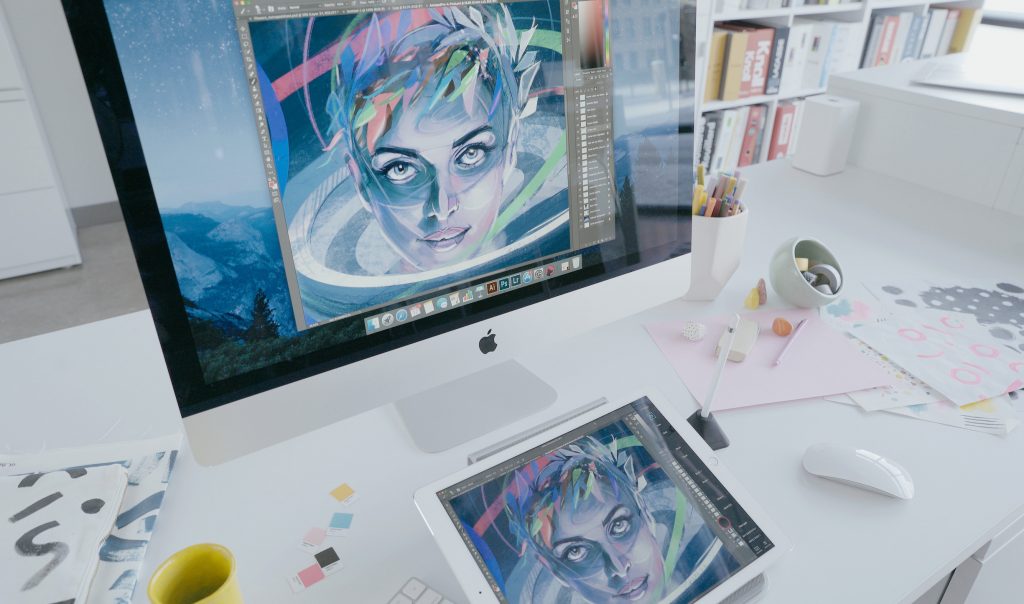 A messy desk with a tablet and computer both displaying a colorful digital drawing of a women's face. 