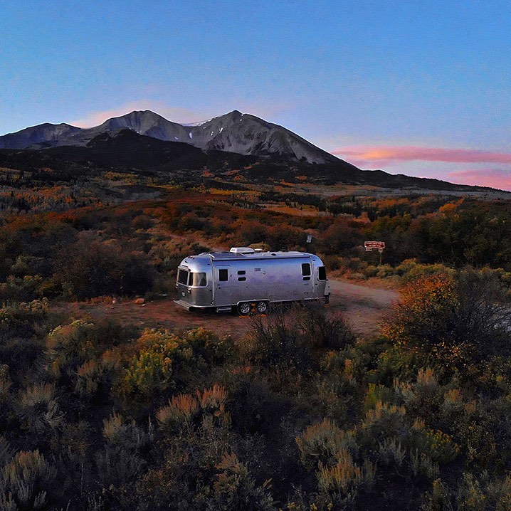 An airstream RV surrounded by bushes in the desert with a mountain in the distance. 