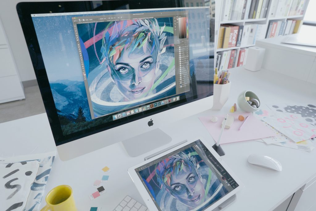 A tablet and computer with a digital drawing of a woman's face on the desk of a cluttered office