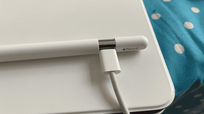 The Apple Pencil USB-C with a USB-C cable plugged into it resting on top of an iPad in a white case. The devices are on blue and white polka-dot fabric