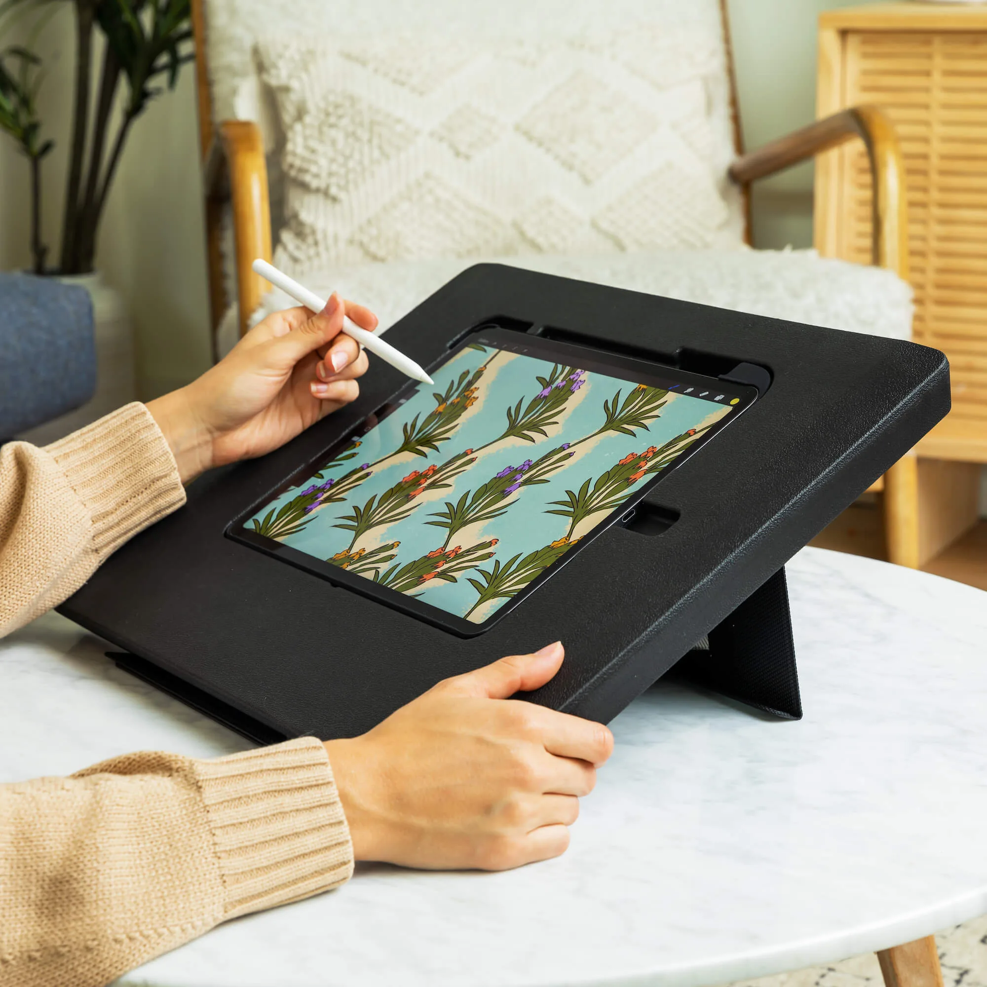 Hands holding a stylus are drawing on a tablet with a floral pattern screen. The tablet is mounted on a black stand, situated on a white table in a cozy living room.