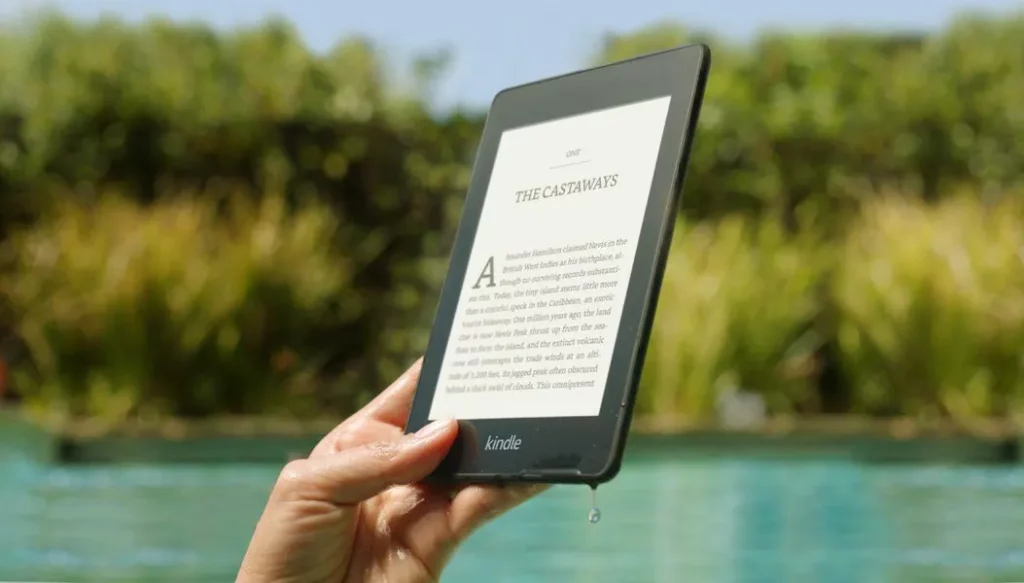 holding up a kindle with a book title called "the castaways". greenery in the background of the image.