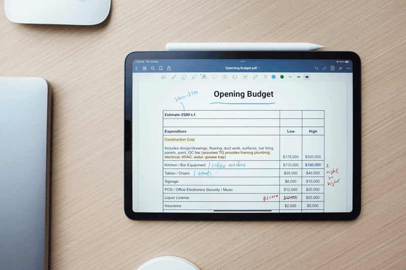 an iPad surrounded by other devices on a wooden desk displaying an annotated "Opening Budget" table in an note-taking app.