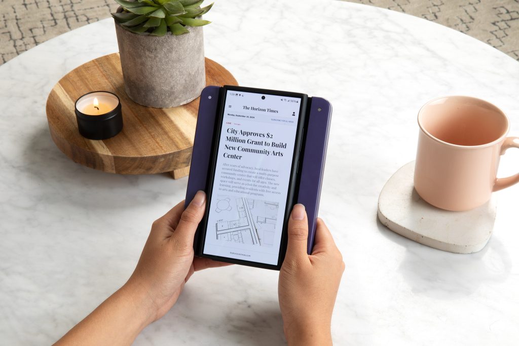 Hands holding Bookcase with android phone at a coffee table. A candle burning and pink coffee cup in background.