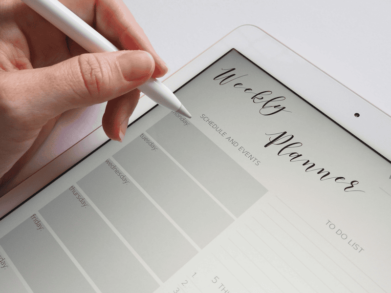 A hand using a stylus to write on a digital weekly planner displayed on an iPad.